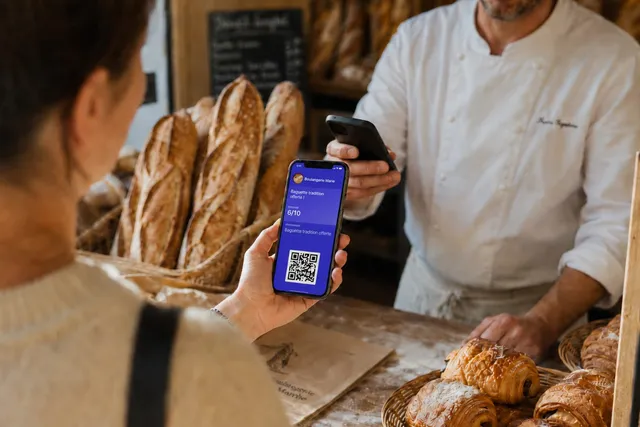 Cliente présentant sa carte de fidélité digitale sur son téléphone au boulanger, qui la scanne devant les baguettes et les viennoiseries